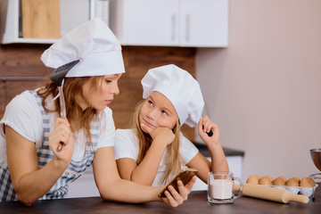 portrait of woman with kid girl thinking about baking something in kitchen, wearing aprons and caps for cooking