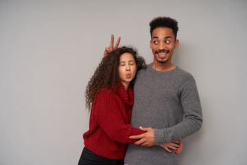 Studio shot of young pretty dark haired curly pair in knitted sweaters having fun together while posing over grey background, embracing each other dearly