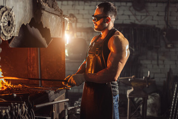 bearded man in good physical form with strong muscles warming iron for manufacturing it, wearing leather apron and protective eyeglasses isolated in workshop