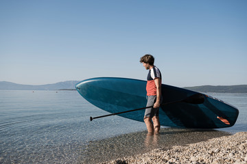 Young man carrying his stand up paddle board into the calm sea water