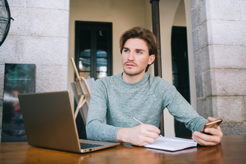 Young freelancer writing in notebook and using smartphone while looking away