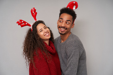 Close-up of joyful young dark haired curly couple dressed in woolen sweaters and christmas hoops enjoying party masquerade and smiling widely while posing over grey background