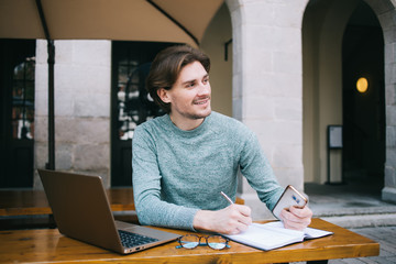 Satisfied hipster man writing in planner while using laptop and sitting at table at street