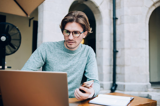 Concentrated hipster man browsing laptop and using smartphone while working in cafe