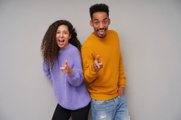 Studio photo of cheerful young dark skinned curly pair pointing happily at camera with raised forefingers and smiling widely, standing over grey background in casual clothes