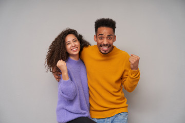 Obraz premium Young happy dark curly brunette couple wearing colourful sweaters while posing over grey background, looking excitedly at camera and smiling joyfully with raised fists
