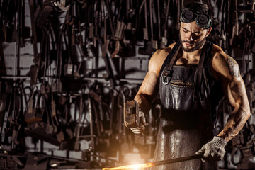 young muscular blacksmith man manually forging the molten metal. Blacksmith hammering hot metal arrow blade, wearing leather apron - Powered by Adobe