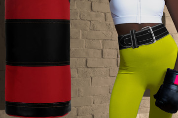 Closeup of black woman in sportswear, boxer glove and powerlifting belt on white brick wall with hanging punching bag background. Concept of gym fitness or sport club visiting and active lifestyle