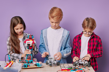 Portrait of children in casual wear, creative teamwork of caucasian kids assembling robots isolated over purple background. science, technique concept