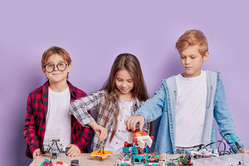 portrait of enthusiastic three kids working as team, assembling robots. three future engineers look at table full of robotics, two boys and one girl