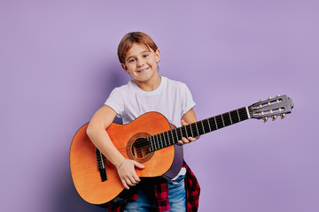 portrait of handsome joyful kid playing on guitar, performing music isolated over purple background