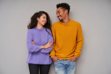 Fototapeta premium Studio photo of charming young curly long haired lady keeping hands folded on her chest while looking happily on her dark skinned cheerful beared boyfriend, isolated over grey background