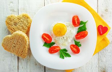 Fried egg and a couple of slices of bread in the shape of a heart. Egg, bread, tomatoes in a white plate on a white wooden background.