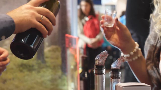 Alcoholic drink tasting at a local indoor market in Lithuania. Woman's hand with bead bracelet holding disposable sampling plastic glass, blurred people in the background.