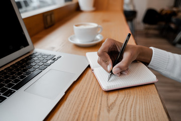 Writer make some notes in notebook with pen, using laptop computer in local cafe and drinking coffee. Digital technology concept and freelancer job remotely. Lifestyle people concept