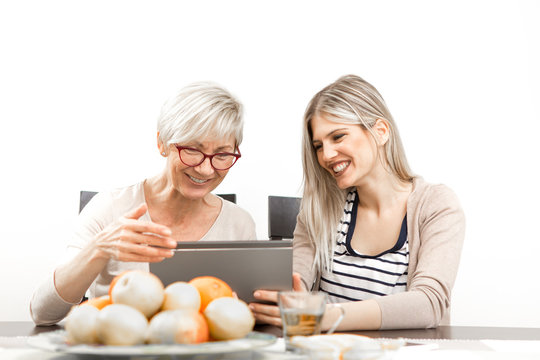 A Senior Blonde Woman Is Sitting In The Dining Room With A Young Woman At The Table. In Front Of Them Is A Tablet Computer, And A Young Woman Is Teaching A Senior Woman How To Use The Interne