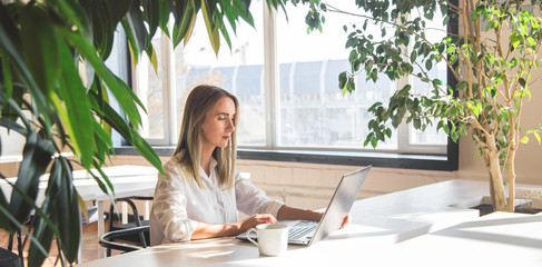 Beautiful caucasian girl working on a laptop remotely in a bright space with green plants.
