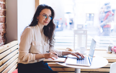 Pleased elegant lady looking away while sitting at table with notepad and laptop in cafe