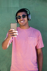 Handsome young guy of color, taking a selfie with headphones, with the pink shirt and in the background a green wall.