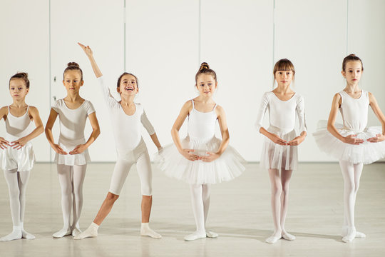 sweet caucasian little girls ballerinas stand in a row in a position wearing white tutu suit, while one girl standing in funny pose, smile, happy ballet group of young girls
