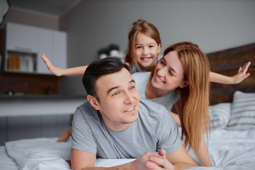 friendly and happy caucasian family, mother and father with child girl lying on bed, wearing casual clothes at home. portrait