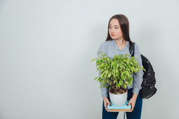 Funny asain student girl standing, holding two books with a flowerpot on the top of them, looking to the side. Backpack slung over her shoulder.