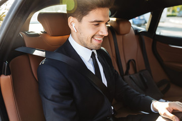 Photo of smiling businessman using earpod and laptop while going in car