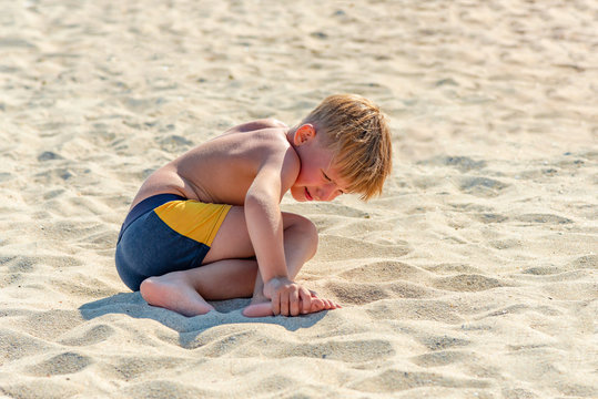 The boy on the beach during the game hit his leg and sits on the sand holding on to it.