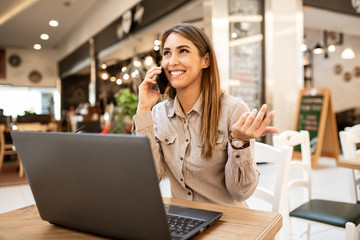 Cute young caucasian businesswoman with brown hair taking a break from work on her laptop and talking on a smart phone with a friend. She is sitting in a cafe.