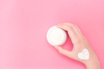 Obraz na płótnie Canvas Hands of a beautiful well-groomed woman with a cream jar on a pink textural background. Moisturizer for clean and soft skin in the winter. The heart shape is made of cream. copyspace. Place for text