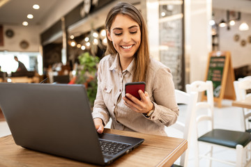Attractive young caucasian businesswoman with brown hair smiling and looking at her smart phone while she is working on her laptop. She is sitting in a coffee shop.