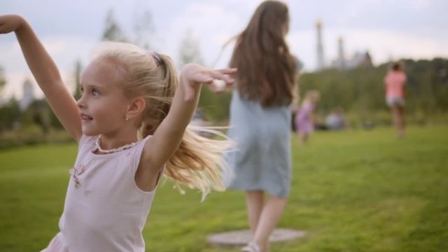 Two charming long-hair sisters 9 and 5 years old in blue and rose dresses dancing on meadow in Moscow downtown. Kremlin and other people are in defocus on a background. Slow motion, cloudy summer day