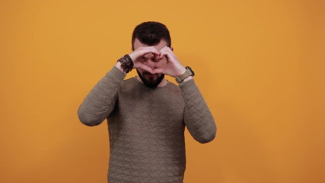 Unhappy handsome caucasian man over isolated orange background doing shape of heart, looking angry wearing casual green clothes