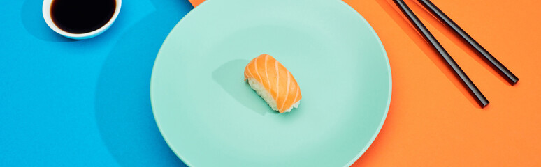 fresh nigiri with salmon near soy sauce and chopsticks on blue and orange surface, panoramic shot