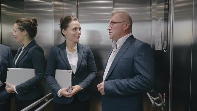 Confident Caucasian Business Colleagues, Man And Woman Exiting Modern Steel Elevator Discussing Their Project At Right Floor