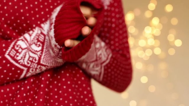 young woman wearing red and white christmas sweater rubbing hands together with christmas lights as a background