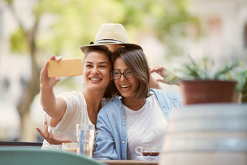 Two gorgeous caucsaian girls sitting in cafe outside and taking selfie.