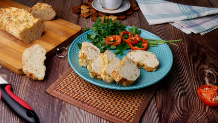 Sliced homemade bread baguette with cheese on top on a white plate with vegetables a wooden table.