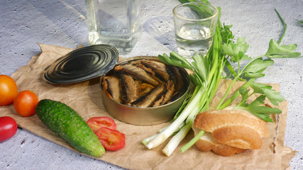 Open can of sprats on paper on a cement background next to bread, vegetables and vodka
