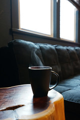 A mug of hot coffee on wooden stool table with cushion sofa on the back