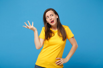 Pensive puzzled young brunette woman girl in yellow t-shirt posing isolated on bright blue wall background, studio portrait. People lifestyle concept. Mock up copy space. Looking up, spreading hands.