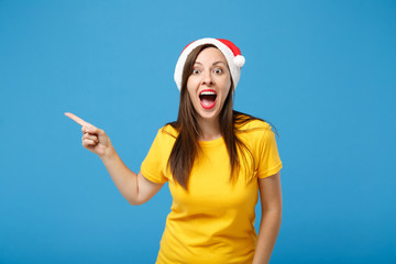 Fototapeta premium Shocked young brunette woman Santa girl in yellow t-shirt Christmas hat posing isolated on blue background. New Year 2020 celebration holiday concept. Mock up copy space. Pointing index finger aside.