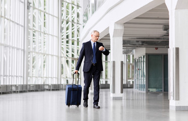 business trip, corporate and people concept - senior businessman walking with travel bag along office building or airport and checking time