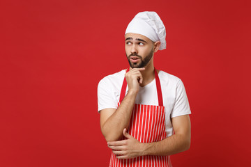 Pensive male chef cook baker man in striped apron white t-shirt toque chefs hat posing isolated on red background. Cooking food concept. Mock up copy space. Put hand prop up on chin, looking aside.