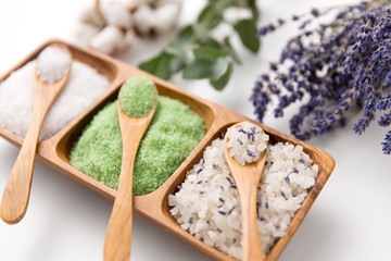 beauty, bath and wellness concept - sea salt with lavender, eucalyptus cinerea, cotton flowers and spoons on wooden tray