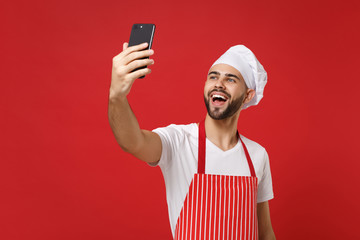 Cheerful young bearded male chef cook or baker man in striped apron toque chefs hat isolated on bright red wall background. Cooking food concept. Mock up copy space. Doing selfie shot on mobile phone.