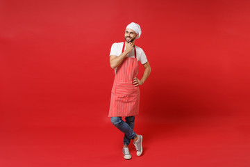 Smiling young bearded male chef cook or baker man in striped apron white t-shirt toque chefs hat posing isolated on red background. Cooking food concept. Mock up copy space. Put hand prop up on chin.
