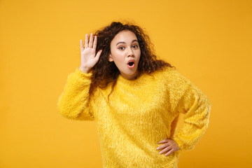 Shocked young african american girl in fur sweater posing isolated on yellow orange background. People lifestyle concept. Mock up copy space. Try to hear you with hand near ear, keeping mouth open.