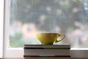 Tea in a yellow cup and stack of books on a rustic window sill. Selective focus, view of garden in the background.
