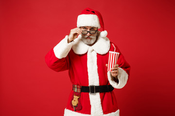 Fototapeta na wymiar Elderly gray-haired mustache bearded Santa man in Christmas hat 3d imax glasses isolated on red background. New Year 2020 celebration concept. Mock up copy space. Watching movie film hold cup of soda.